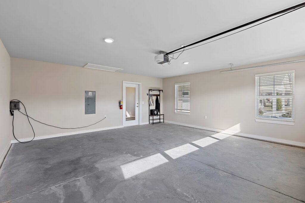 Empty two-car garage with concrete floor, beige walls, two windows, a door, electrical panel, wall-mounted device, and ceiling garage door opener.