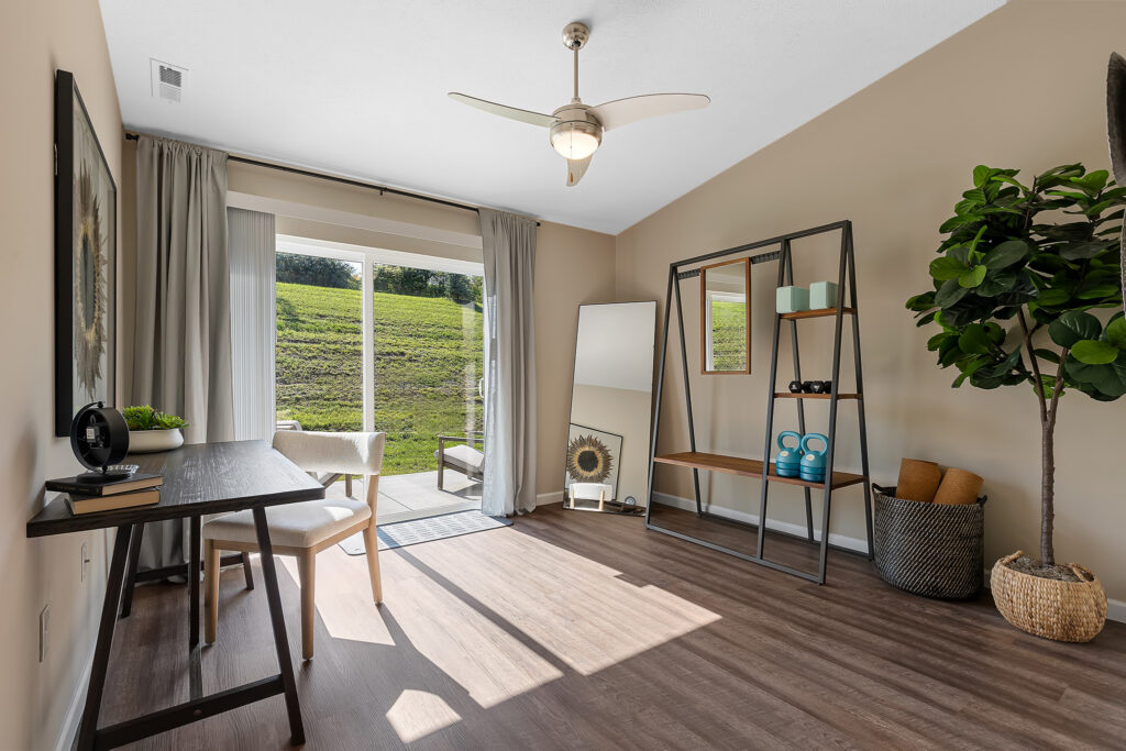 Bright home office with a desk, chair, large shelving unit, mirror, potted plant, and sliding glass door overlooking a grassy hill.