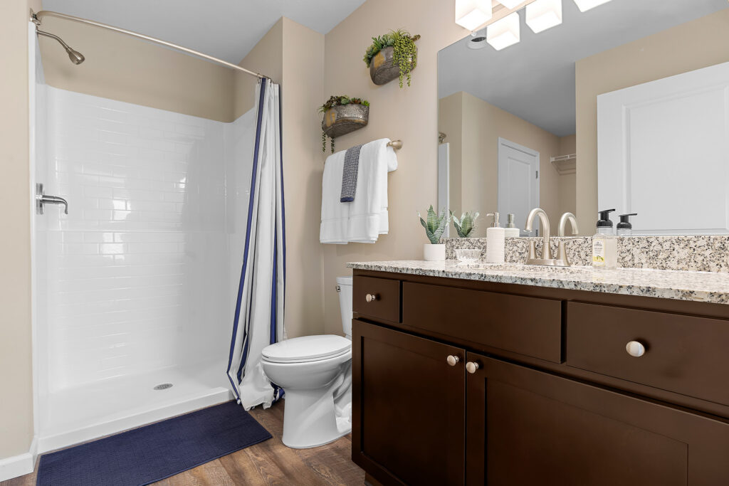 Modern bathroom with a glassless shower, white toilet, granite countertop, dark wood vanity, and neutral walls. Towels and plants decorate the space.