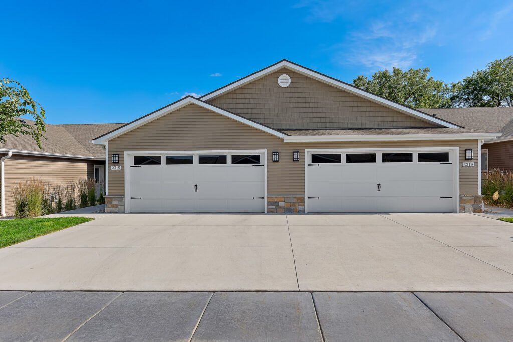 A suburban duplex with two double garages, beige siding, white trim, stone accents, and a wide concrete driveway under a clear blue sky.