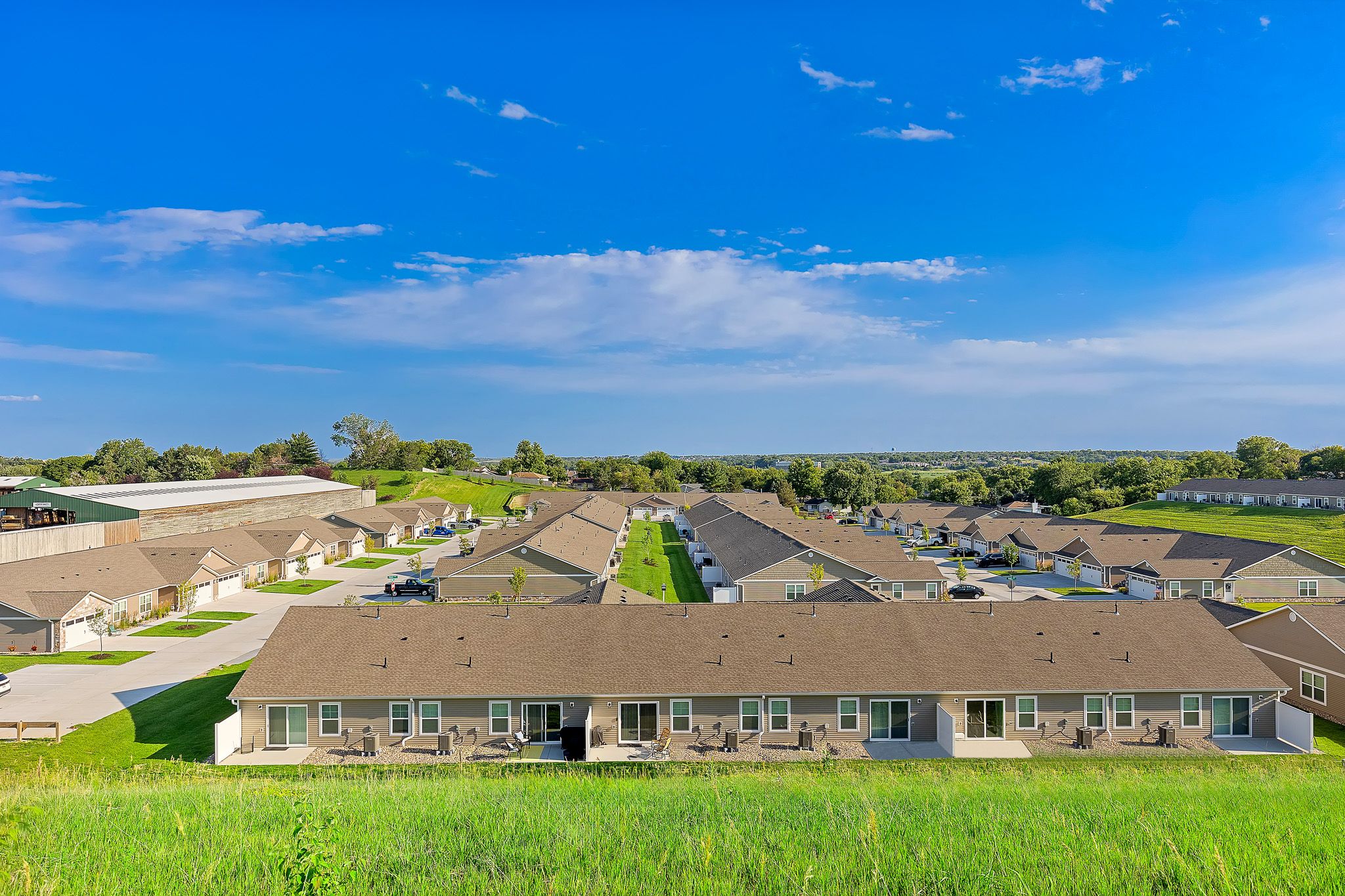 A row of single-story townhomes with porches faces a green lawn under a blue sky, with more similar buildings and trees in the background.