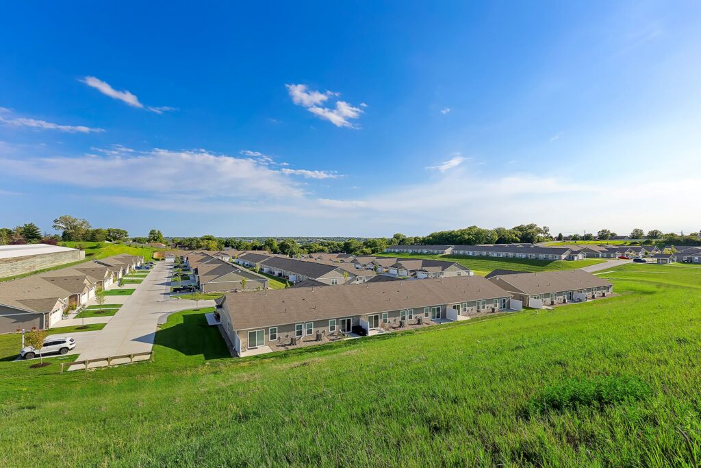 Rows of single-story townhomes with driveways are set in a suburban neighborhood, bordered by green grass and trees under a clear blue sky.