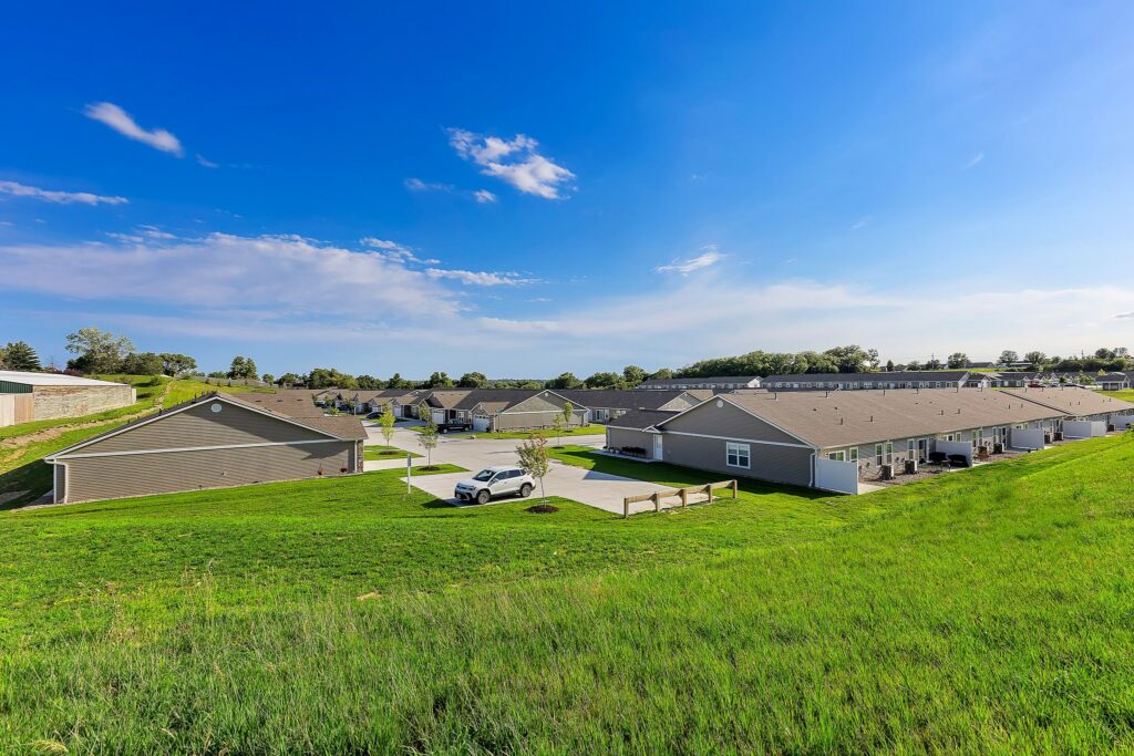 A row of single-story gray residential buildings sits on a gentle grassy slope under a bright blue sky with scattered clouds.