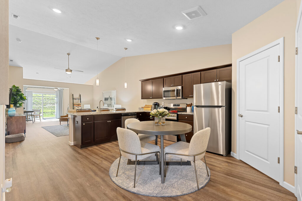 Open kitchen and dining area with dark wood cabinets, stainless steel appliances, round table with four chairs, and view of living room and backyard through glass doors.