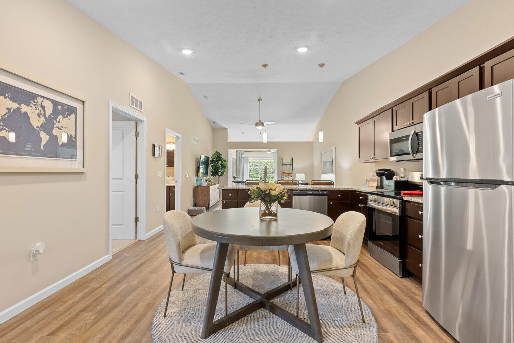 Open kitchen and dining area with a round table, four chairs, dark cabinets, stainless steel appliances, wood flooring, and neutral wall colors.