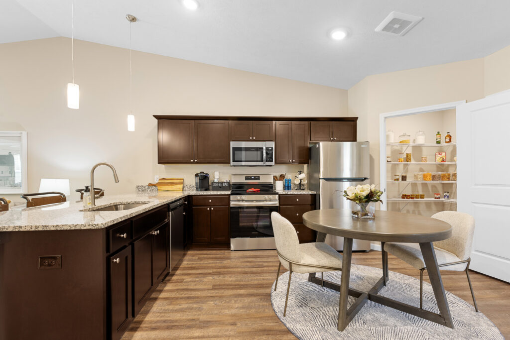 Modern kitchen with dark wood cabinets, stainless steel appliances, island sink, round dining table with two chairs, and an open pantry stocked with food items.