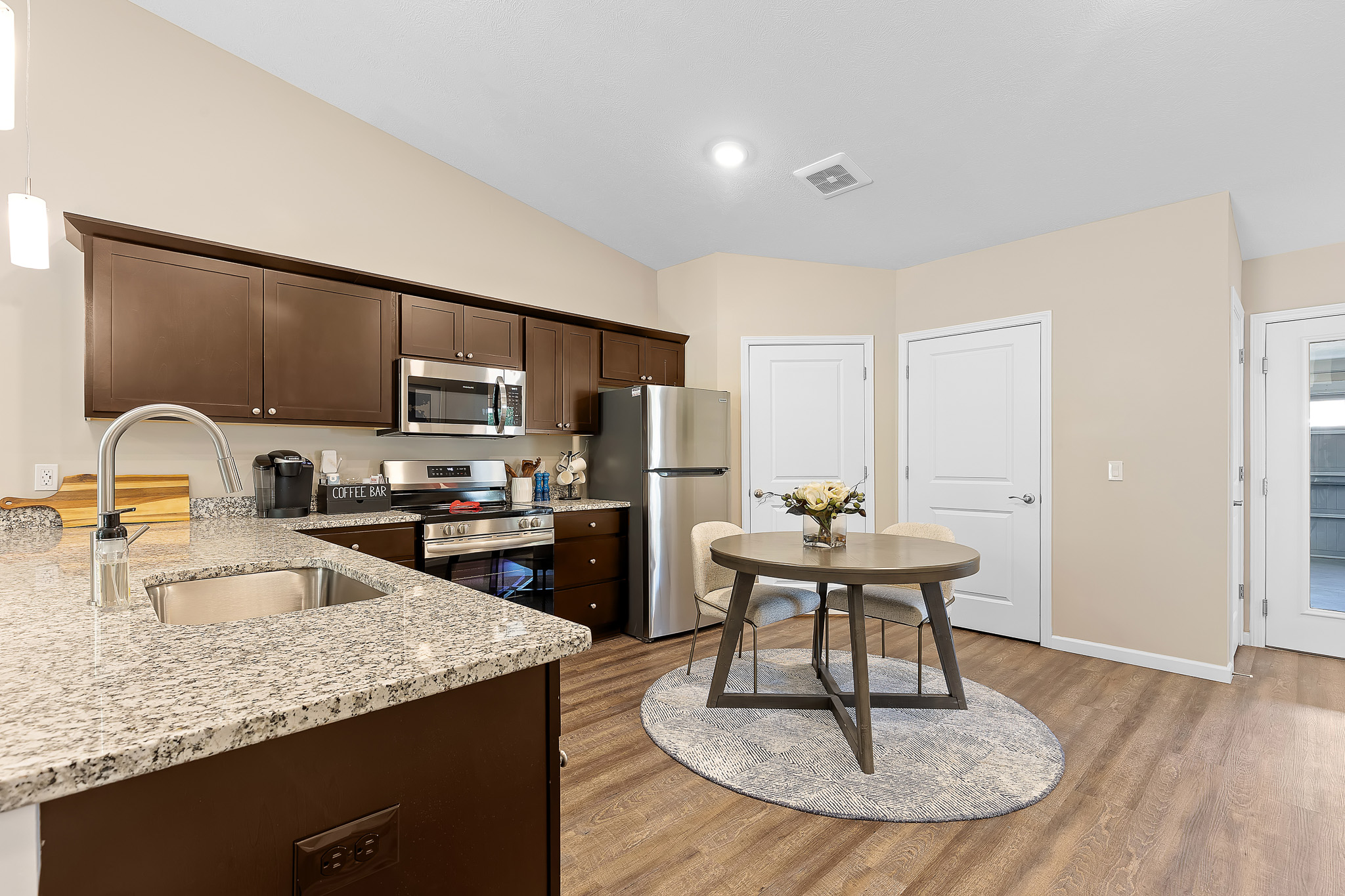 Modern kitchen with granite countertops, stainless steel appliances, dark wood cabinets, round dining table on a rug, and wood flooring.