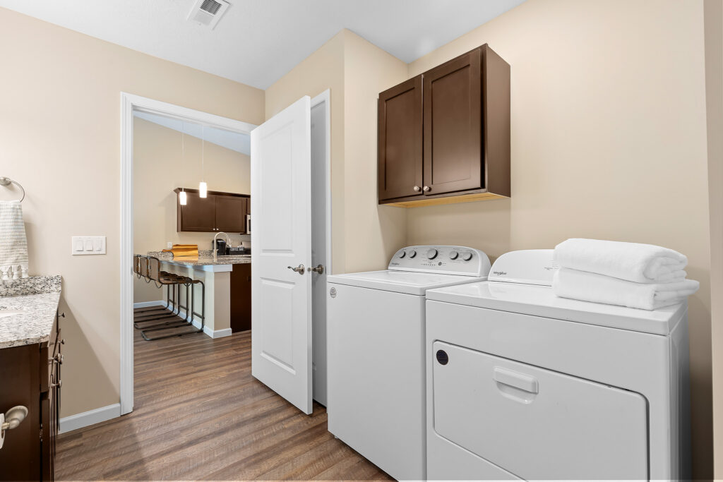 Laundry room with a washer, dryer, folded towels on top, brown cabinets above, and a view into a kitchen with barstools in the background.