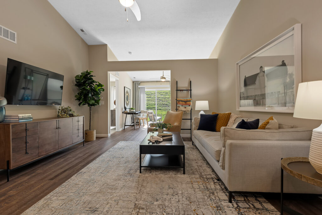 Modern living room with a beige sofa, large wall art, coffee table, TV on a wood console, and view into a dining area with plants. Neutral tones and soft lighting create a cozy atmosphere.