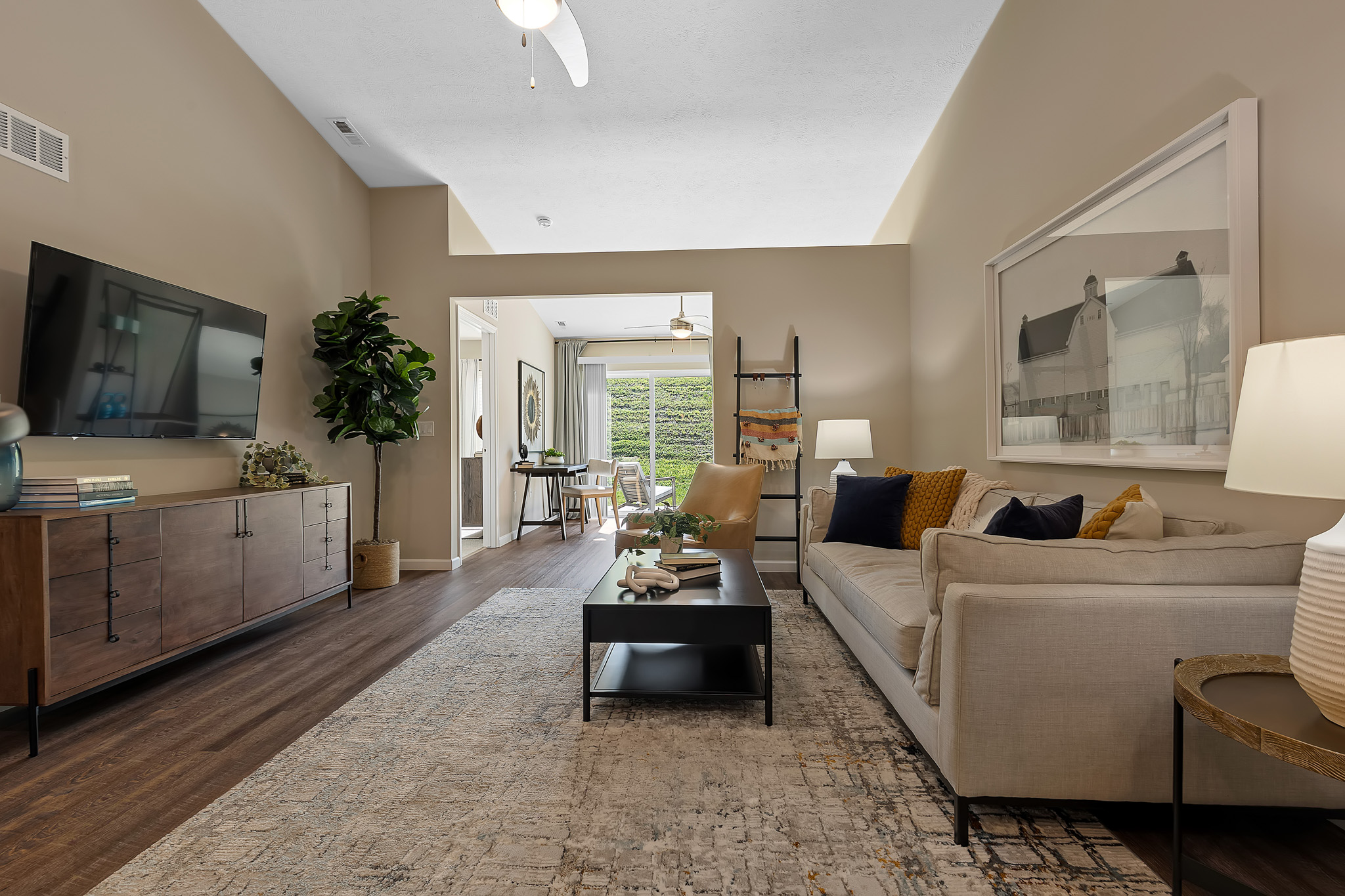 Modern living room with a beige sofa, large wall art, coffee table, TV on a wood console, and view into a dining area with plants. Neutral tones and soft lighting create a cozy atmosphere.