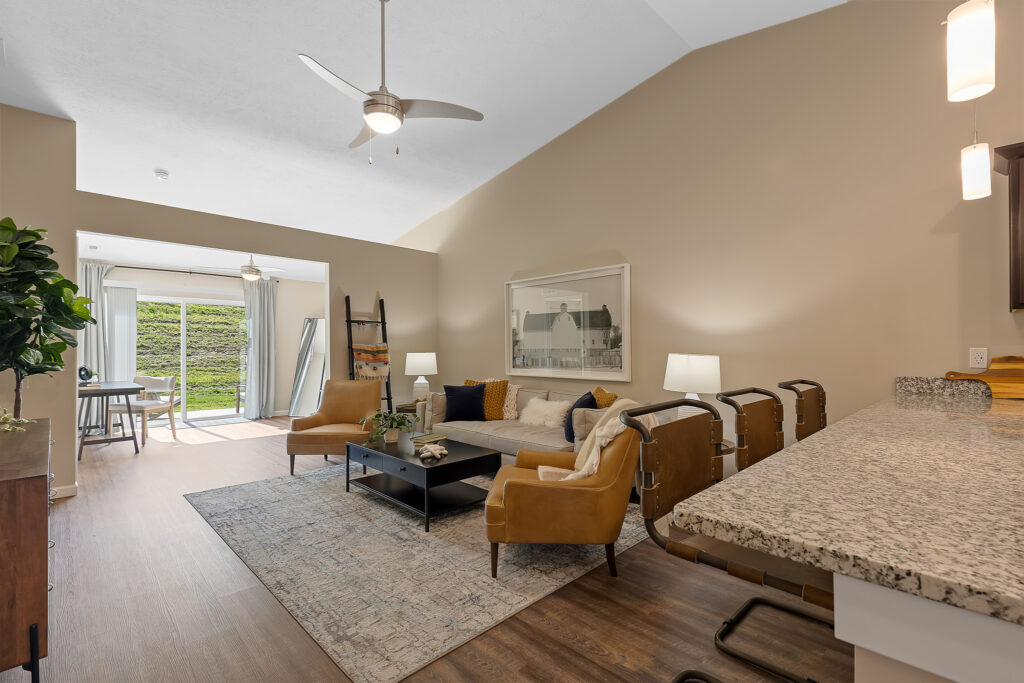 Modern living room with tan walls, brown furniture, a light area rug, and a granite kitchen counter; large windows let in natural light and offer a view of greenery outside.