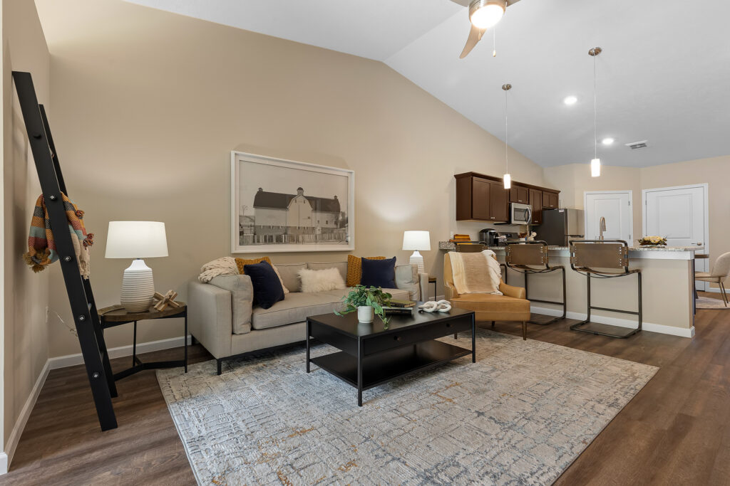 Modern open living room with beige walls, a gray sofa, black coffee table, kitchen island with barstools, pendant lights, and wooden flooring. Artwork and decor create a cozy atmosphere.