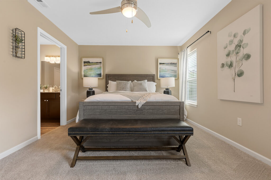 A neatly made bed with two nightstands, a black bench at the foot, beige walls, and framed art in a bright, carpeted bedroom.