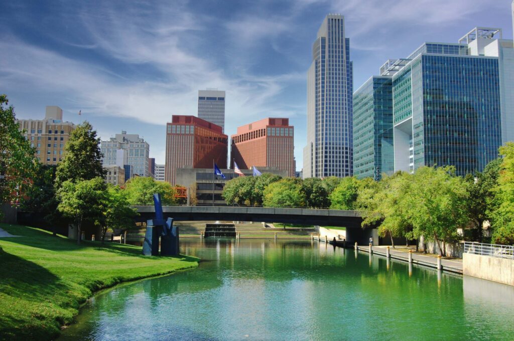 A cityscape with modern office buildings behind a river lined with grass and trees, under a partly cloudy sky.