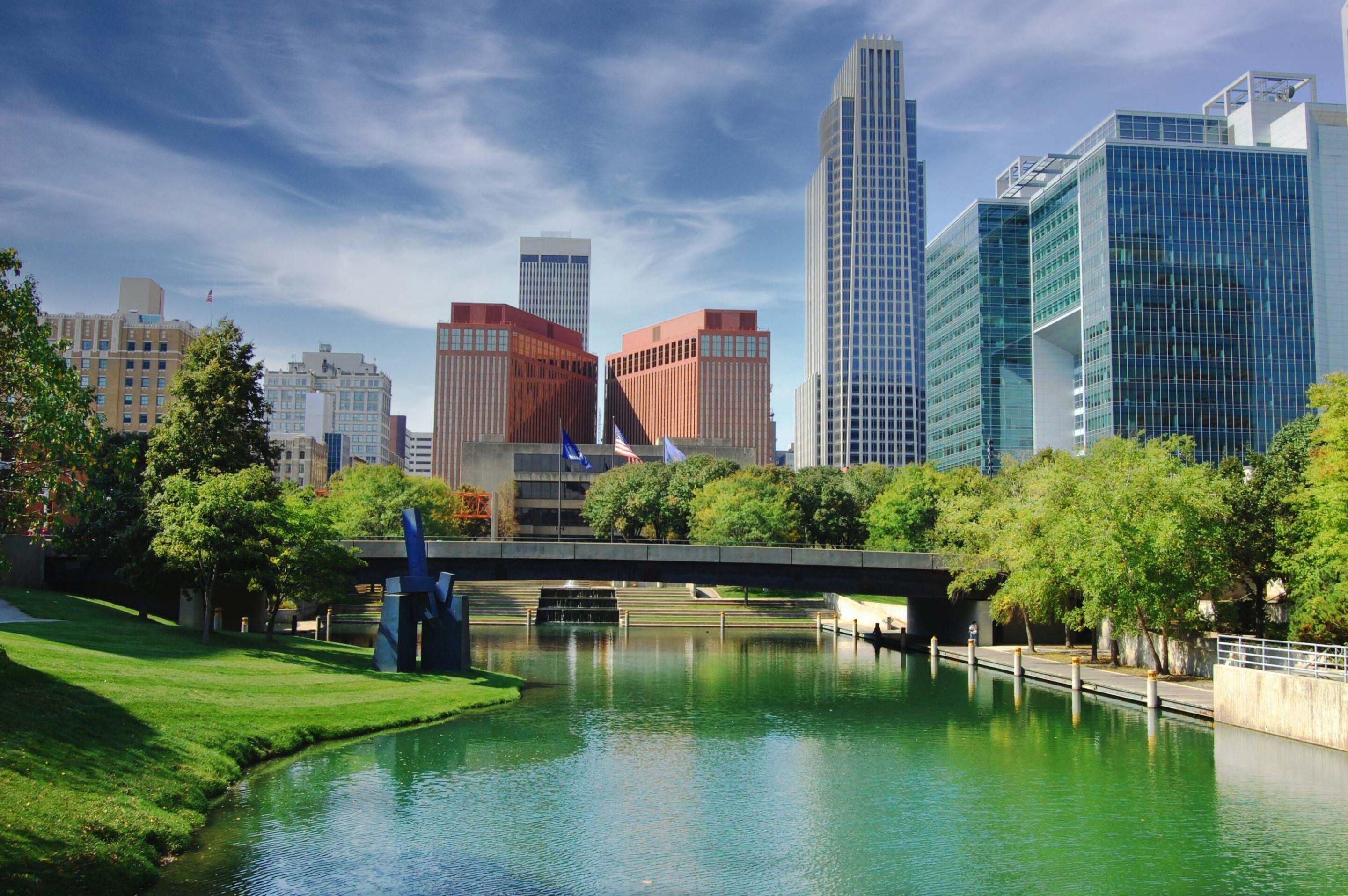 A cityscape with modern office buildings behind a river lined with grass and trees, under a partly cloudy sky.