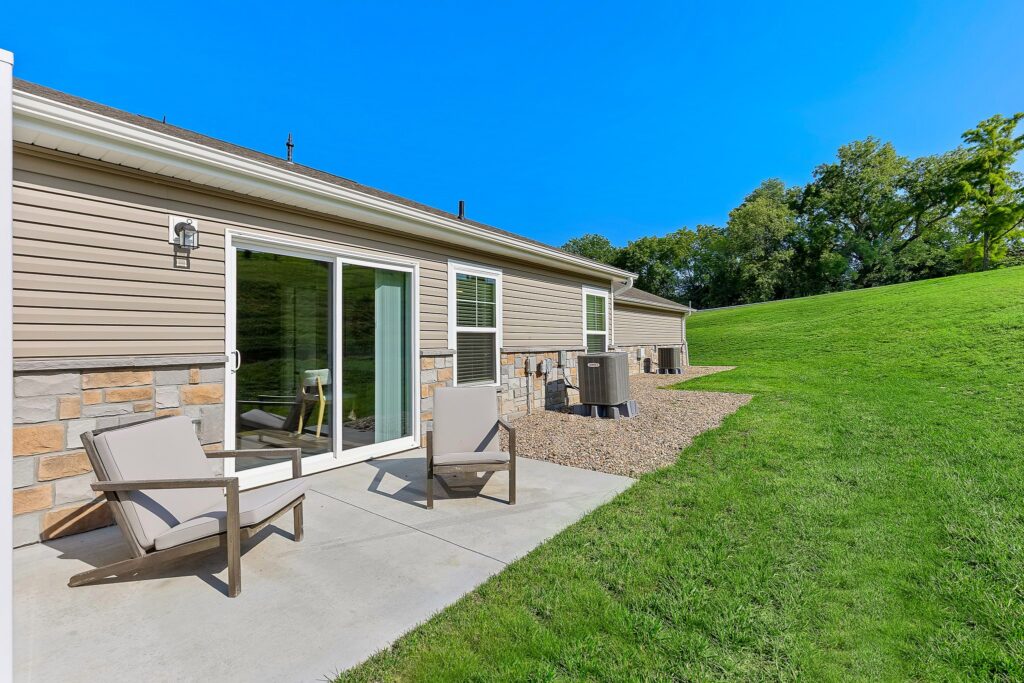 A concrete patio with two chairs behind a single-story house, beige siding, and sliding glass doors, next to a grassy lawn and a line of trees.