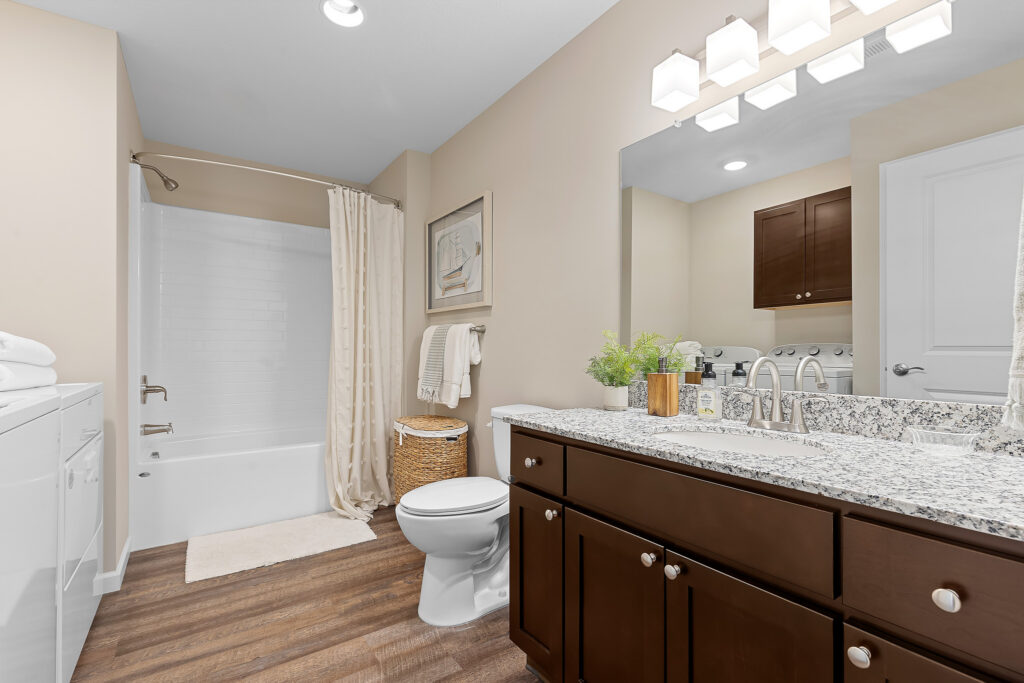 Modern bathroom with a granite countertop, dark wood cabinets, white toilet and bathtub, laundry machines, and neutral decor.