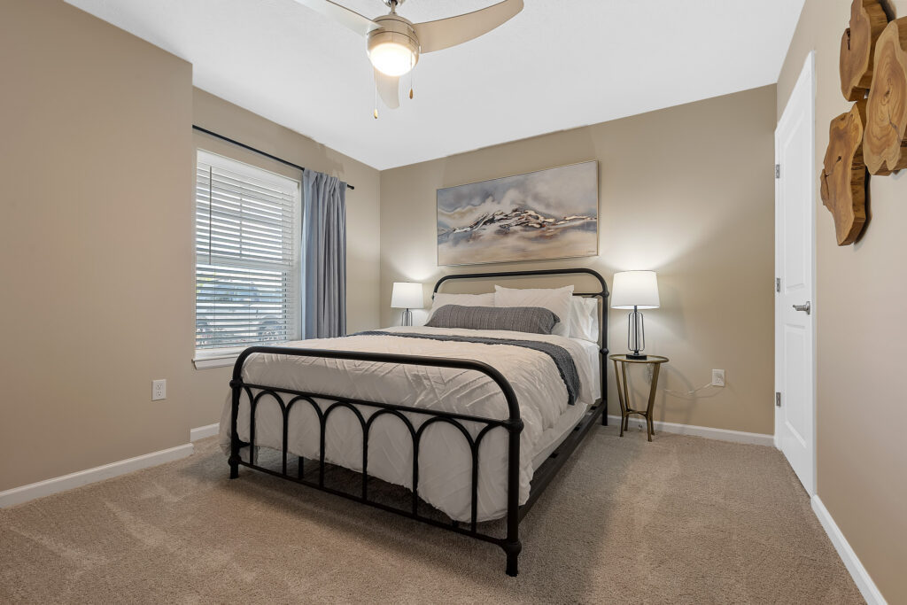 A neatly made bed with white and gray bedding sits between two lamps in a beige carpeted bedroom with a window, artwork, and ceiling fan.
