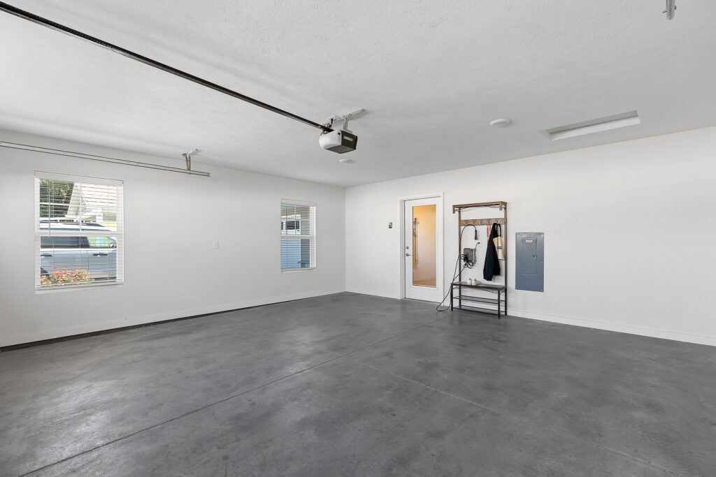 Empty garage with concrete floor, white walls, two windows, a door, a metal rack with hanging items, and a ceiling-mounted garage door opener.