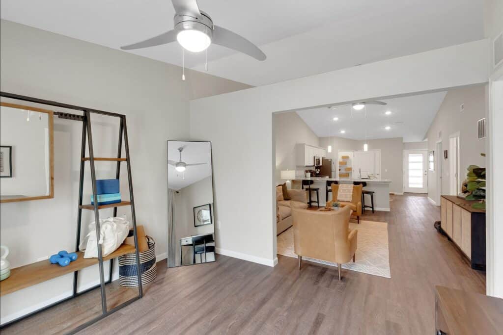 Open living space with wood flooring, modern furnishings, ceiling fans, and neutral decor; view includes a dining area, kitchen, and seating area with tan chairs and a standing mirror.