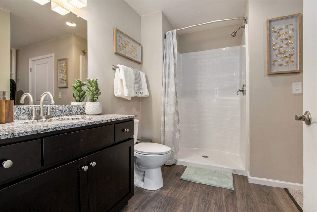 Modern bathroom with dark vanity, granite countertop, toilet, walk-in shower, wall art, towel rack, and potted plant on wood-style flooring.