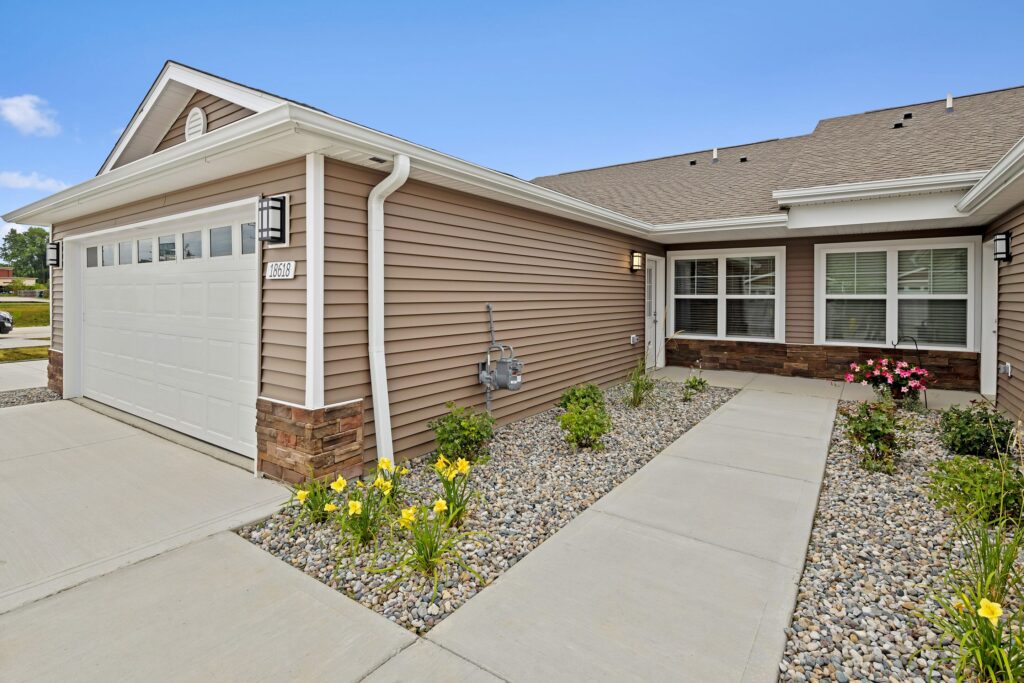 Single-story house with beige siding, a two-car garage, flower beds with yellow and pink blooms, and a concrete walkway leading to the front door.
