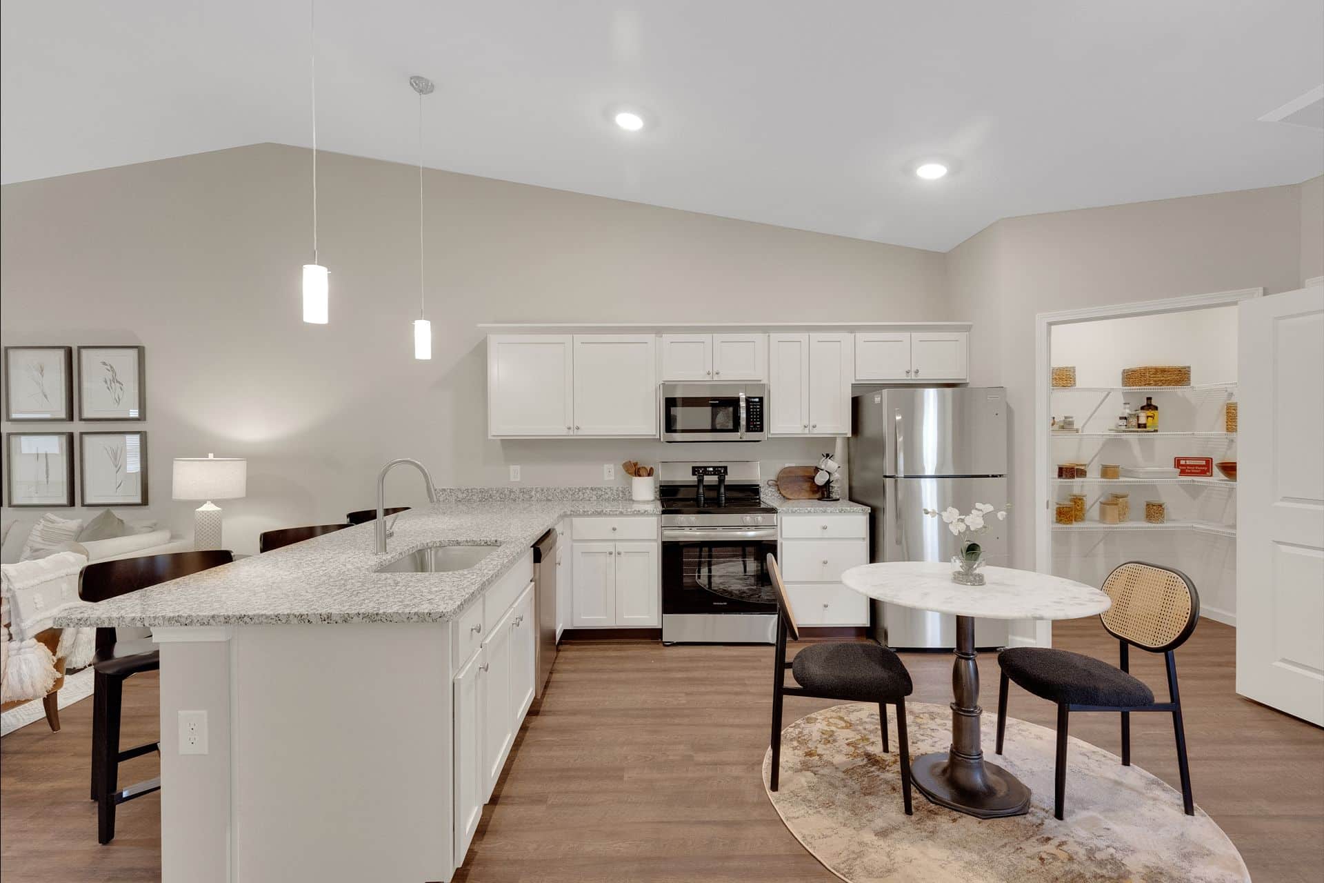 Modern kitchen with white cabinets, stainless steel appliances, an island with sink and seating, and a small dining table with two chairs on a round rug. The open pantry is visible in the background, adding to the inviting atmosphere of this Redwood Canton Michigan Avenue home.