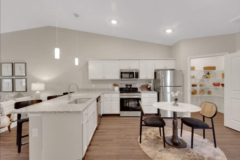 Modern kitchen with white cabinets, stainless steel appliances, a granite island with bar stools, and a small round dining table with two chairs on a rug. Open pantry shelves are visible in the background.
