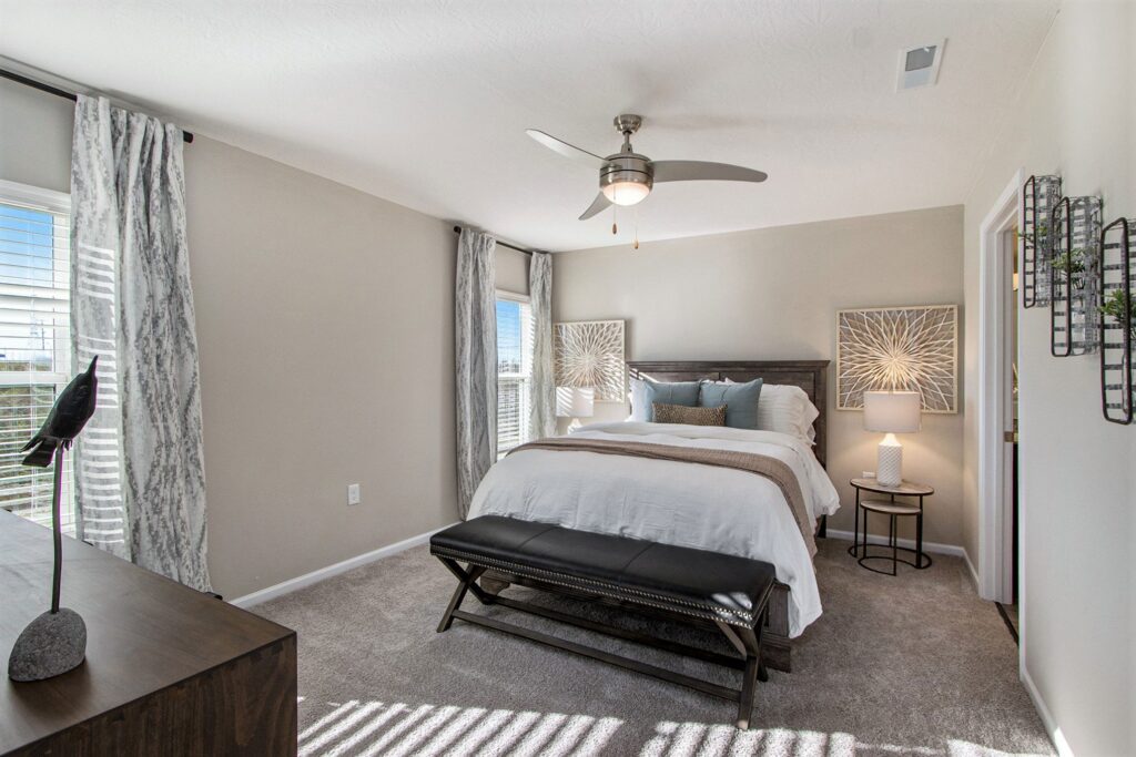 Modern bedroom with a queen bed, neutral decor, ceiling fan, two windows with curtains, a dresser, and a bench at the foot of the bed. Light gray walls and carpeted floor.