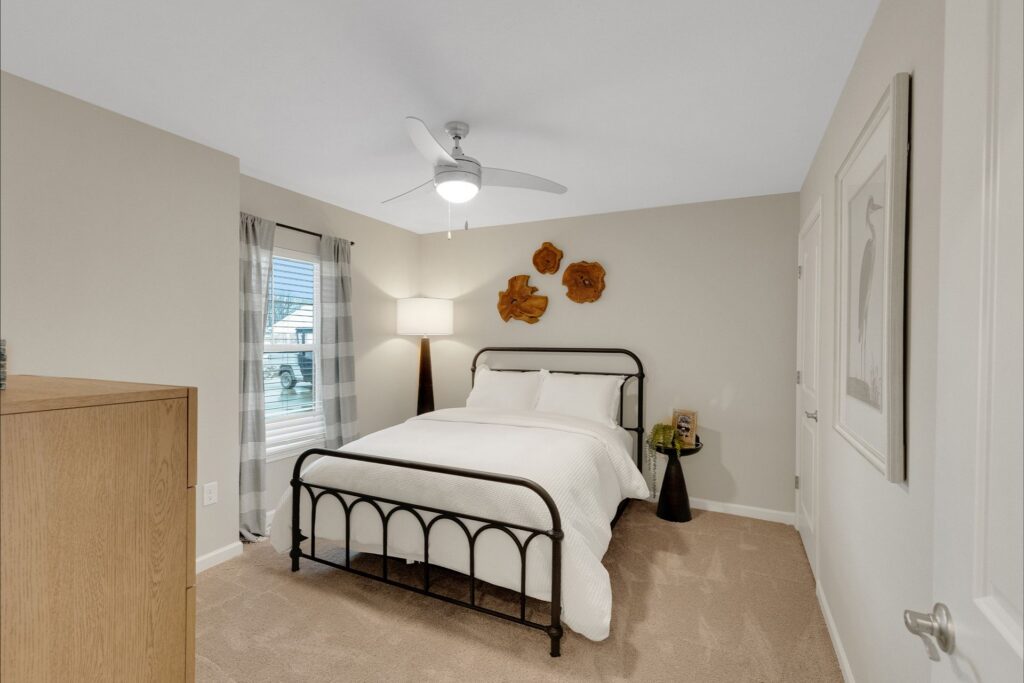 Neutral bedroom with a double bed, white bedding, black metal frame, wall art, side table, and a window with plaid curtains; ceiling fan and lamp provide lighting.