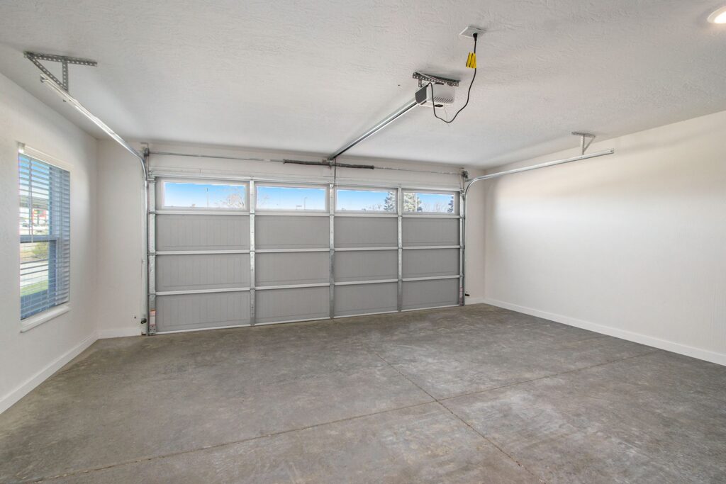 Empty, clean two-car garage with closed paneled metal door, concrete floor, white walls, ceiling opener, and a window on the left letting in natural light.