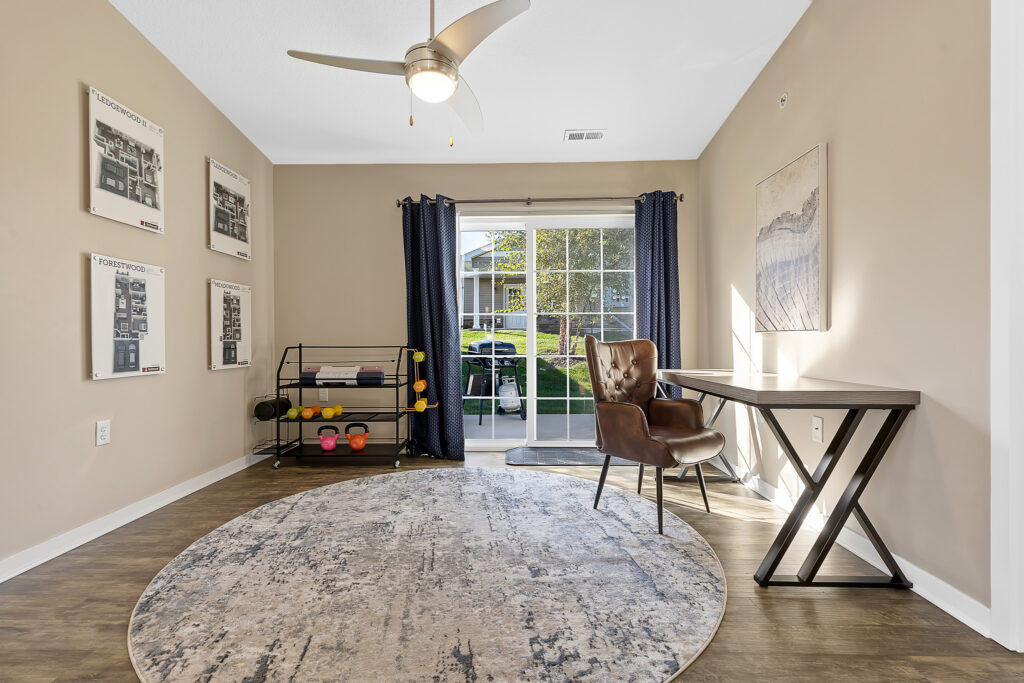 Home office with a desk, chair, and shelving unit for weights. Large window with curtains overlooks a grassy yard. Neutral walls and round rug on wood flooring.