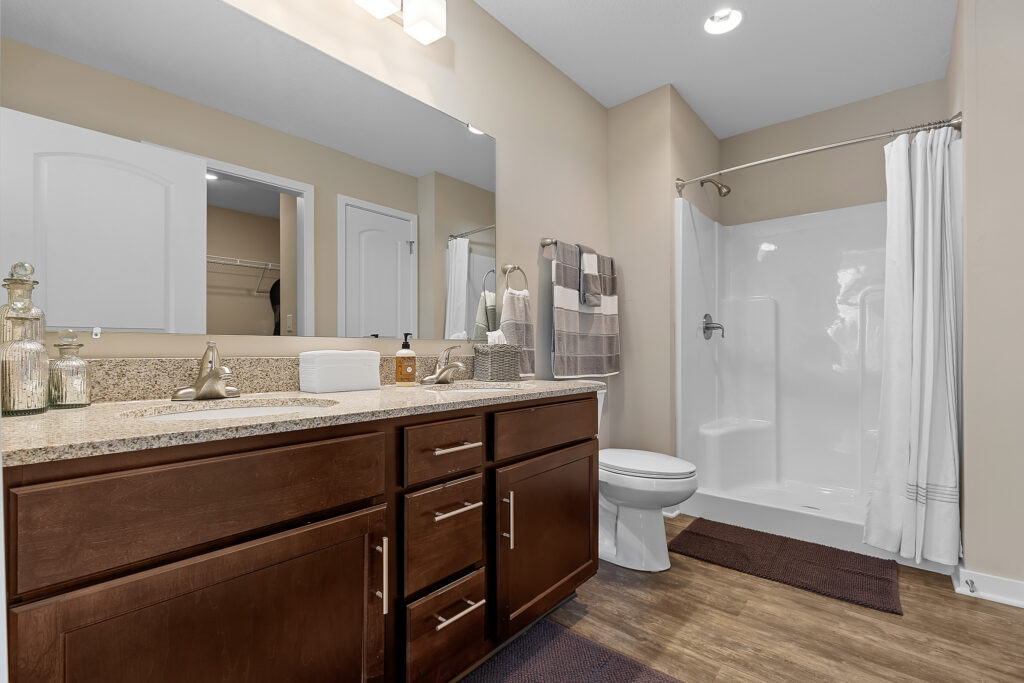 Modern bathroom with double sink vanity, granite countertop, toilet, walk-in shower with white curtain, beige walls, and wood-style flooring. Towels and decor jars are on the counter and rack.