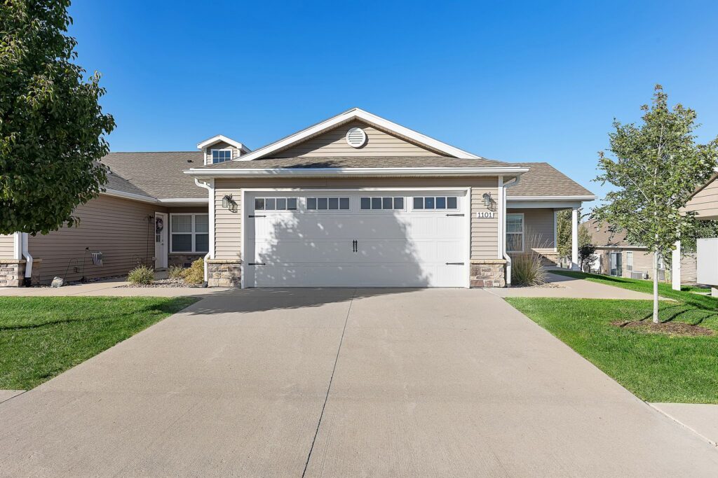 Single-story suburban house with a double garage, beige siding, manicured lawn, and a clear blue sky.