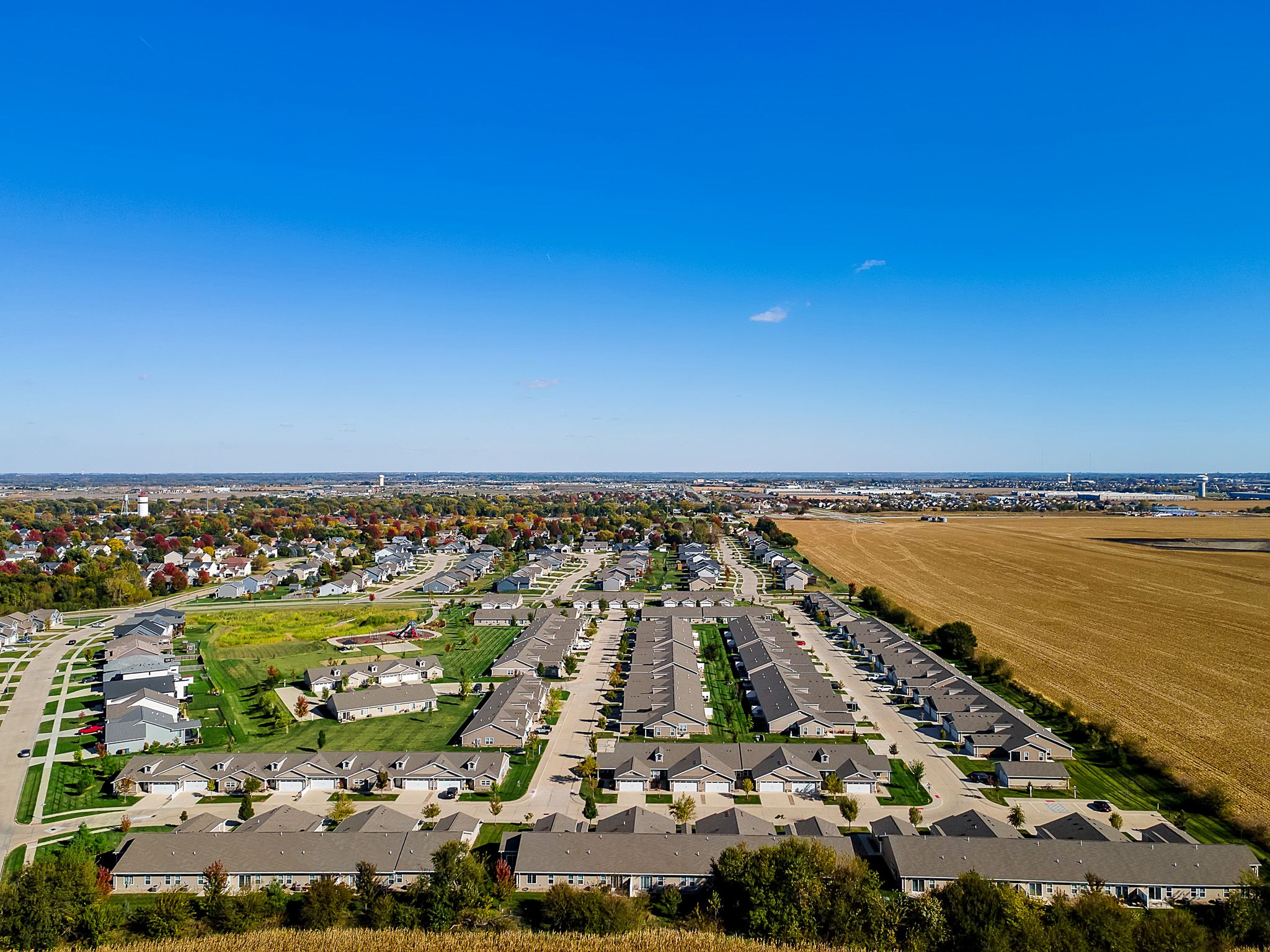 Aerial view of a suburban neighborhood with rows of houses, green lawns, roads, and an adjacent large field under a clear blue sky.