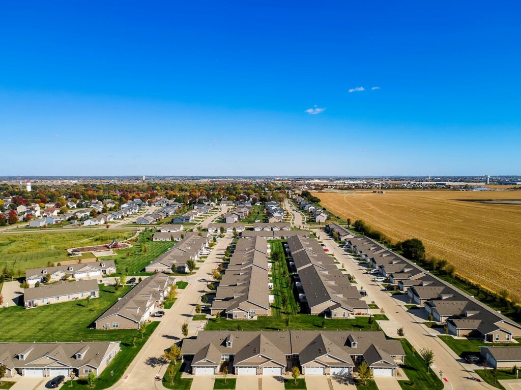 Aerial view of a suburban neighborhood with rows of houses, green lawns, and a large field to the right under a clear blue sky.