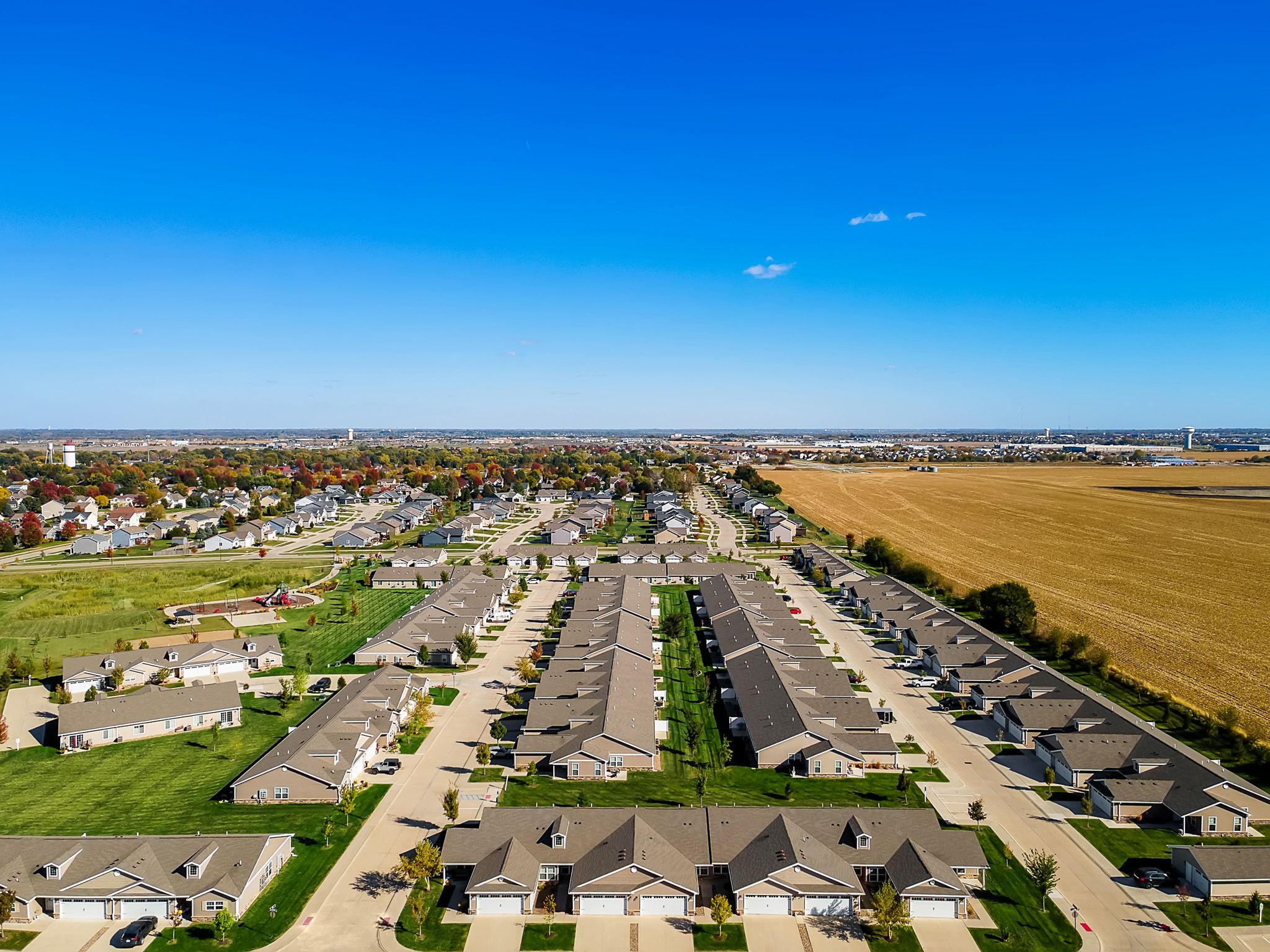 Aerial view of a suburban neighborhood with rows of houses, green lawns, and a large field to the right under a clear blue sky.