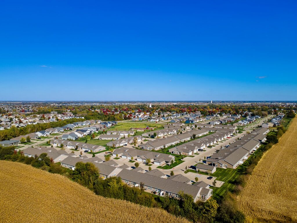 Aerial view of a suburban neighborhood with rows of houses, green lawns, and surrounding fields under a clear blue sky.