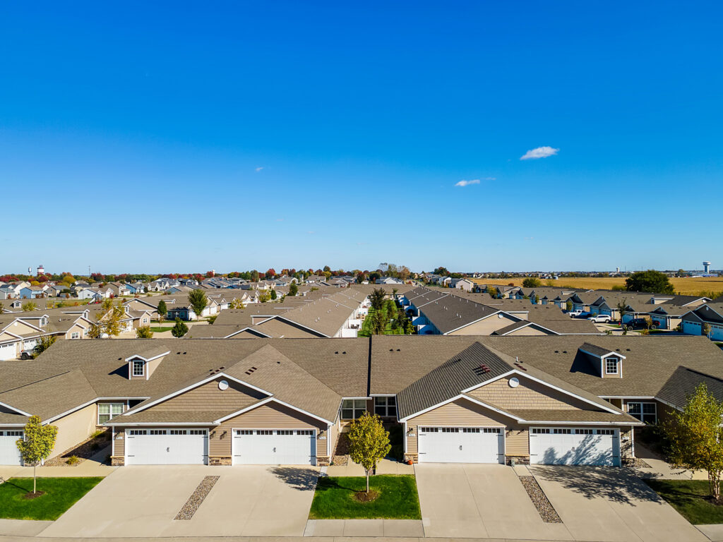 Aerial view of a suburban neighborhood with rows of identical houses, driveways, garages, and neatly trimmed lawns under a clear blue sky.