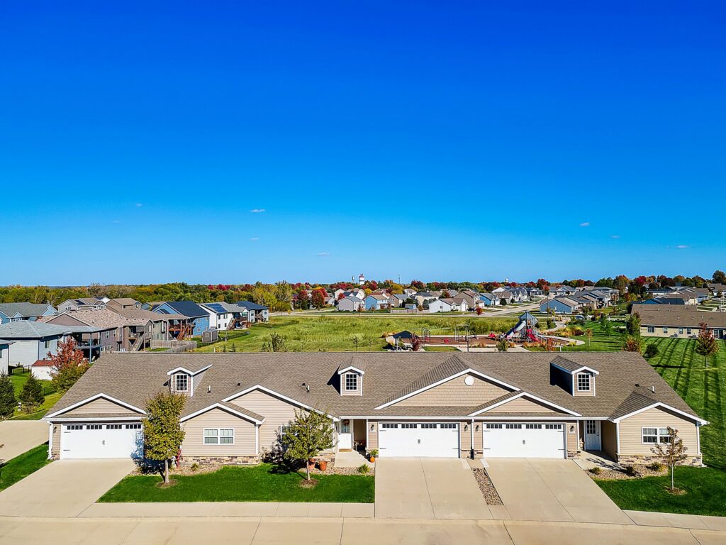 A row of modern townhouses with attached garages under a clear blue sky, surrounded by green lawns and other houses in a suburban neighborhood.