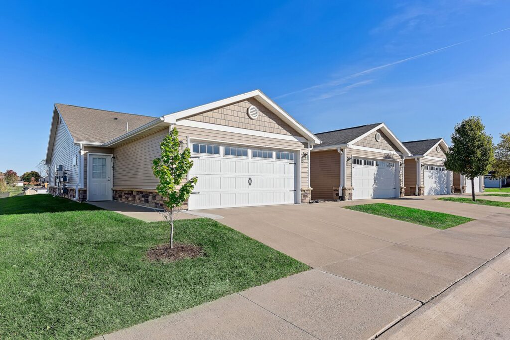 Row of single-story suburban houses with attached garages and driveways, each with well-maintained lawns, under a clear blue sky.