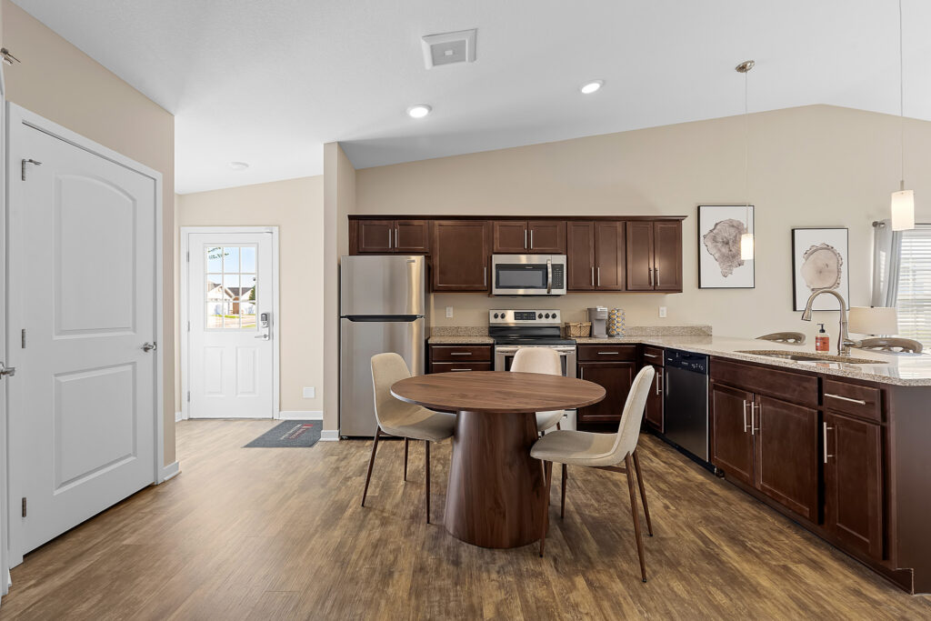 Modern kitchen with dark wood cabinets, stainless steel appliances, round table with four chairs, and wood-style flooring. Two framed prints hang above the counter. Door and window provide natural light.
