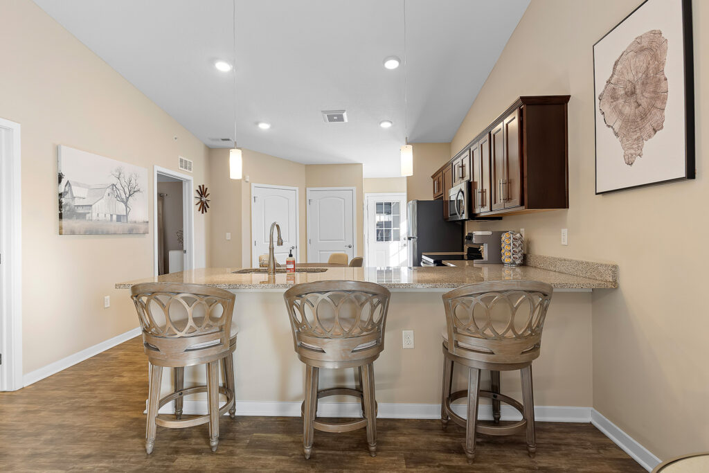 Modern kitchen with three wooden barstools at a granite island, stainless steel appliances, and neutral-colored decor, including framed wall art and pendant lighting.
