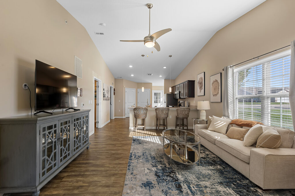 Modern living room with beige walls, a large window, sofa, TV on a console, glass coffee table, bar stools at a kitchen counter, and wood flooring.