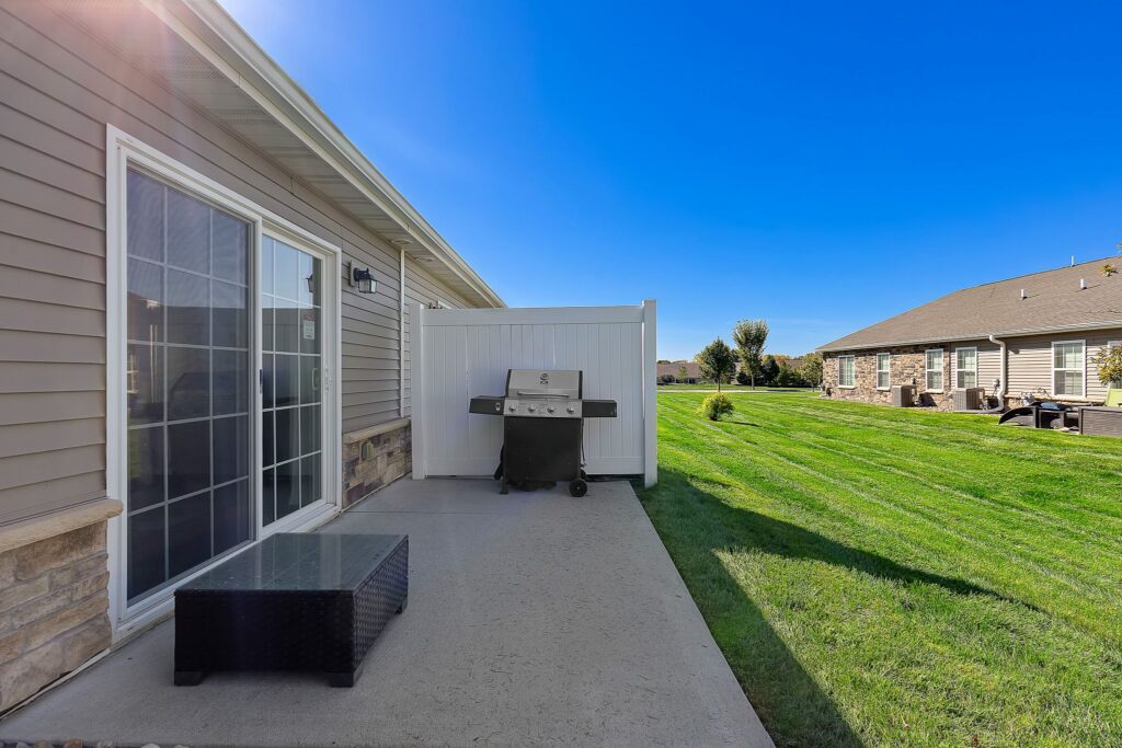 Concrete patio with a glass sliding door, black wicker table, and grill beside a white privacy fence, overlooking a well-maintained grassy yard and neighboring houses under a clear blue sky.