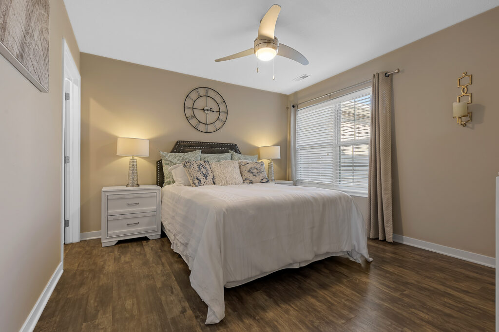 A bedroom with a bed, two nightstands with lamps, a large clock on the wall, a ceiling fan, wooden flooring, and a window with blinds and curtains.