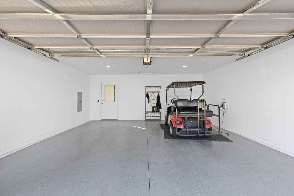 Two-car garage with white walls, gray floor, ceiling lights, a parked golf cart on the right, wall hooks with bags, and a closed door at the back.