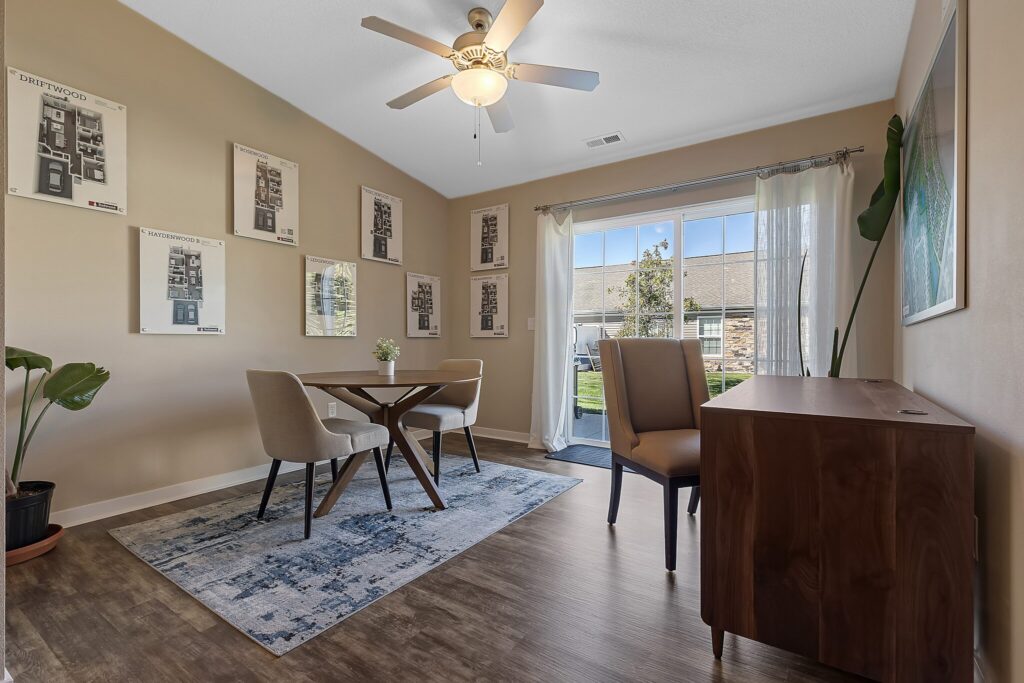A modern home office with a round table, four chairs, a wooden desk, framed art on beige walls, and a glass door leading to a grassy outdoor area.
