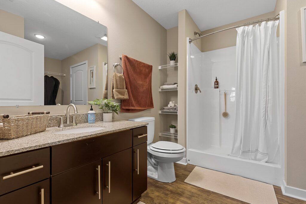 Modern bathroom with a shower, toilet, brown vanity with sink, open shelves holding towels and decor, tan walls, and wood-style flooring.
