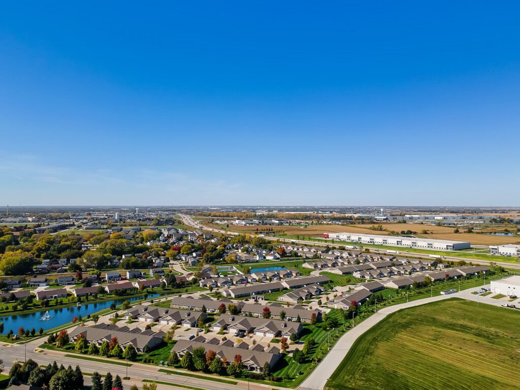 Aerial view of a suburban neighborhood with houses, green lawns, a pond, and roads under a clear blue sky.